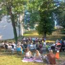 assemblée de personnes disposées en arc de cercle autour d'un orchestre, sur une pelouse, dans un parc entouré d'arbre et au pied du bâtiment d'archives