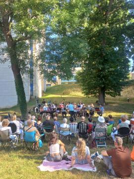 assemblée de personnes disposées en arc de cercle autour d'un orchestre, sur une pelouse, dans un parc entouré d'arbre et au pied du bâtiment d'archives