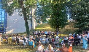 assemblée de personnes disposées en arc de cercle autour d'un orchestre, sur une pelouse, dans un parc entouré d'arbre et au pied du bâtiment d'archives