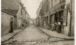 Vue d'une rue, sur la droite à l'entrée de la rue, une boutique "épicerie, mercerie, vins et liqueurs" devant laquelle se trouvent un homme et deux femmes
