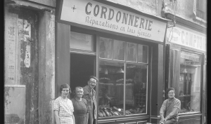 Trois personnes se trouvent sur le seuil d'une boutique de coordonier, une quatrième est devant la boutique, sur un vélo