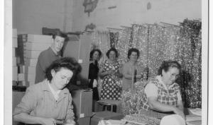 Deux femmes travaillent, penchées sur une table, derrière elles trois femmes et un hommes semblent observer la scène