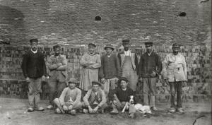 Des ouvriers de la tuilerie en blouses et vestes de travail se tiennent debout devant des tas de briques, au premier rang, trois jeunes ouvriers sont assis et l'un d'eux est accompagné d'un chien couché sur ses genoux