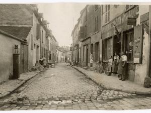 Vue d'une rue, sur la droite à l'entrée de la rue, une boutique "épicerie, mercerie, vins et liqueurs" devant laquelle se trouvent un homme et deux femmes