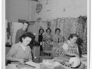 Deux femmes travaillent, penchées sur une table, derrière elles trois femmes et un hommes semblent observer la scène