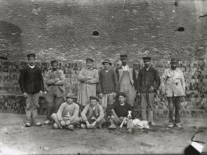 Des ouvriers de la tuilerie en blouses et vestes de travail se tiennent debout devant des tas de briques, au premier rang, trois jeunes ouvriers sont assis et l'un d'eux est accompagné d'un chien couché sur ses genoux