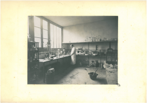 Louis Vernin dans un laboratoire entouré de fioles, flacons, pillons.