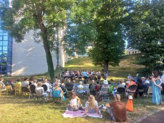 assemblée de personnes disposées en arc de cercle autour d'un orchestre, sur une pelouse, dans un parc entouré d'arbre et au pied du bâtiment d'archives