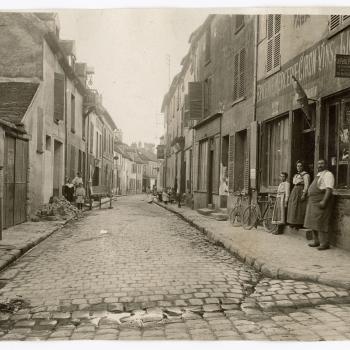 Vue d'une rue, sur la droite à l'entrée de la rue, une boutique "épicerie, mercerie, vins et liqueurs" devant laquelle se trouvent un homme et deux femmes