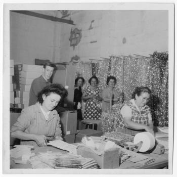 Deux femmes travaillent, penchées sur une table, derrière elles trois femmes et un hommes semblent observer la scène