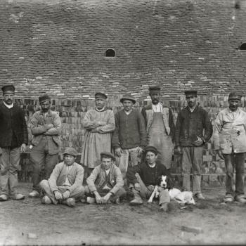 Des ouvriers de la tuilerie en blouses et vestes de travail se tiennent debout devant des tas de briques, au premier rang, trois jeunes ouvriers sont assis et l'un d'eux est accompagné d'un chien couché sur ses genoux