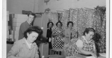 Deux femmes travaillent, penchées sur une table, derrière elles trois femmes et un hommes semblent observer la scène