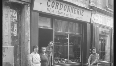 Trois personnes se trouvent sur le seuil d'une boutique de coordonier, une quatrième est devant la boutique, sur un vélo