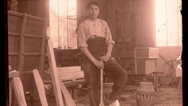 Un homme se tient debout dans un atelier, il s'appuie sur une hâche et l'un de ses pieds est posé sur une pièce de bois.