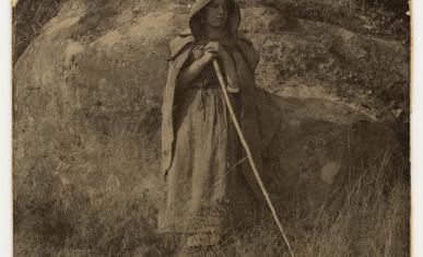 Jeune femme tenant un bâton de marche et portant une pèlerine, adossée à un rocher