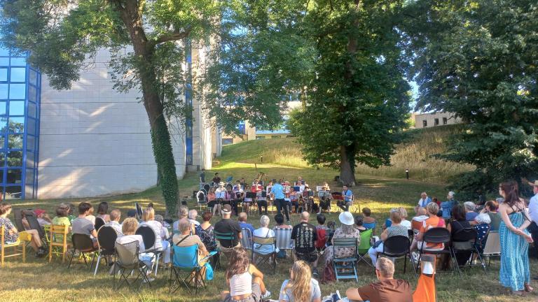 assemblée de personnes disposées en arc de cercle autour d'un orchestre, sur une pelouse, dans un parc entouré d'arbre et au pied du bâtiment d'archives