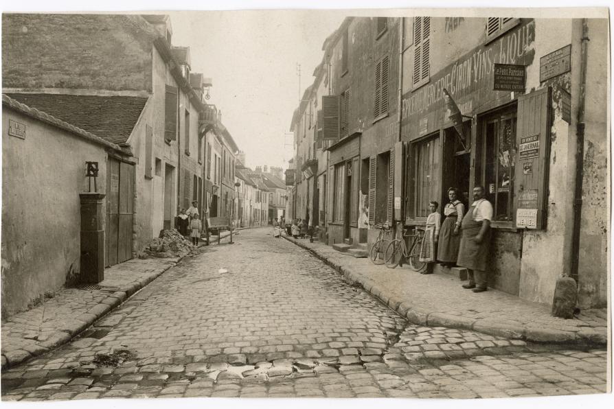 Vue d'une rue, sur la droite à l'entrée de la rue, une boutique "épicerie, mercerie, vins et liqueurs" devant laquelle se trouvent un homme et deux femmes