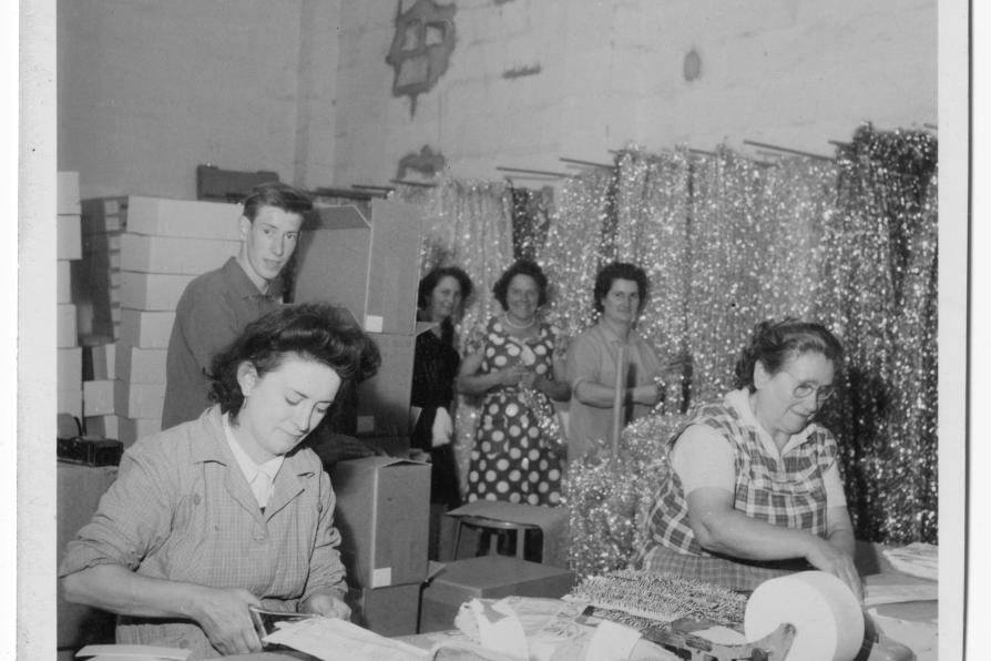 Deux femmes travaillent, penchées sur une table, derrière elles trois femmes et un hommes semblent observer la scène