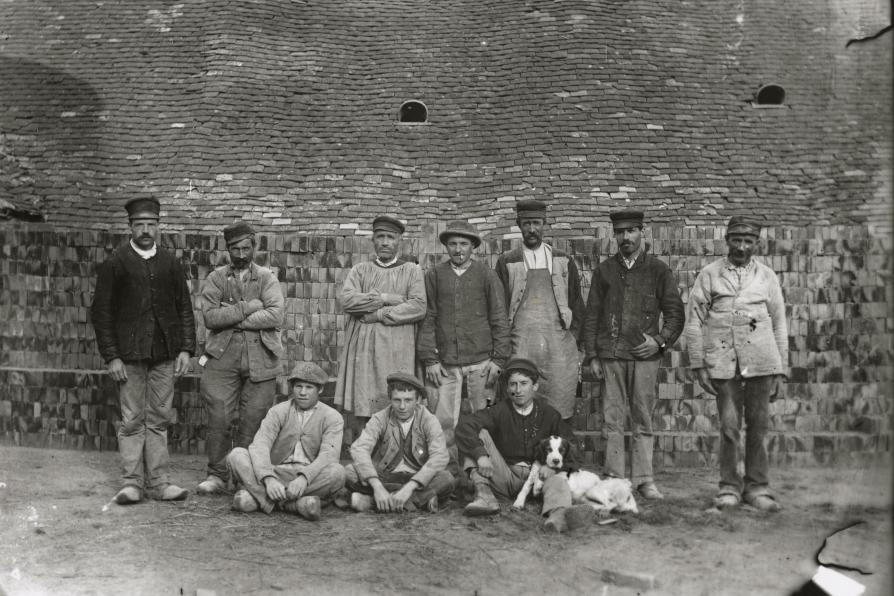Des ouvriers de la tuilerie en blouses et vestes de travail se tiennent debout devant des tas de briques, au premier rang, trois jeunes ouvriers sont assis et l'un d'eux est accompagné d'un chien couché sur ses genoux